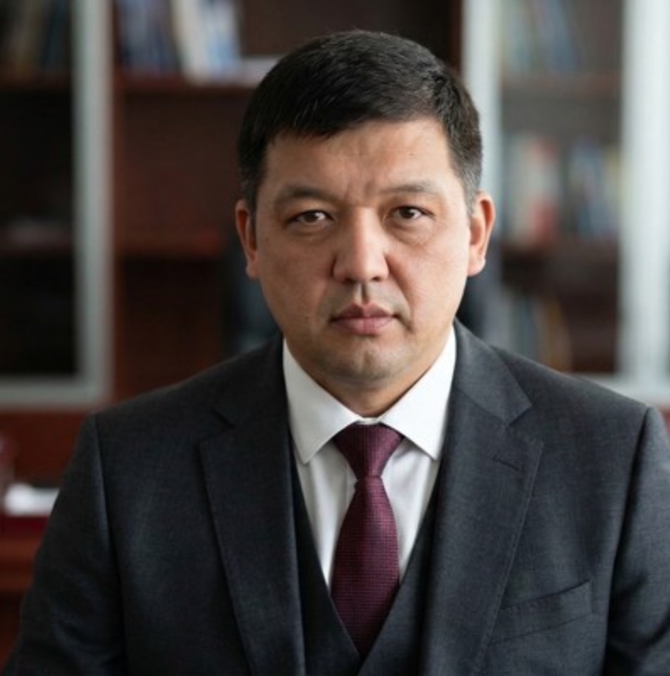 Айдар Абишев в тёмном костюме и бордовом галстуке на фоне офиса.